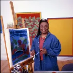 A Black woman in a long denim shirt smiling beside a painting of a landscape on an easel.