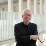 An older man in a black suit jacket and black shirt looks up with a slight smile as he holds black sunglasses in his right hand, with arms crossed. White architecture—many thin columns and a kind of fence stand behind him.