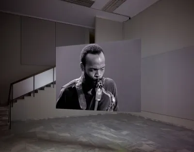 A video projection of a Black man playing a saxophone shown on a hanging screen in an empty gallery.