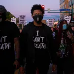 A Black man stands in the center of a color photograph. He wears a face mask and a T-shirt that says "I can