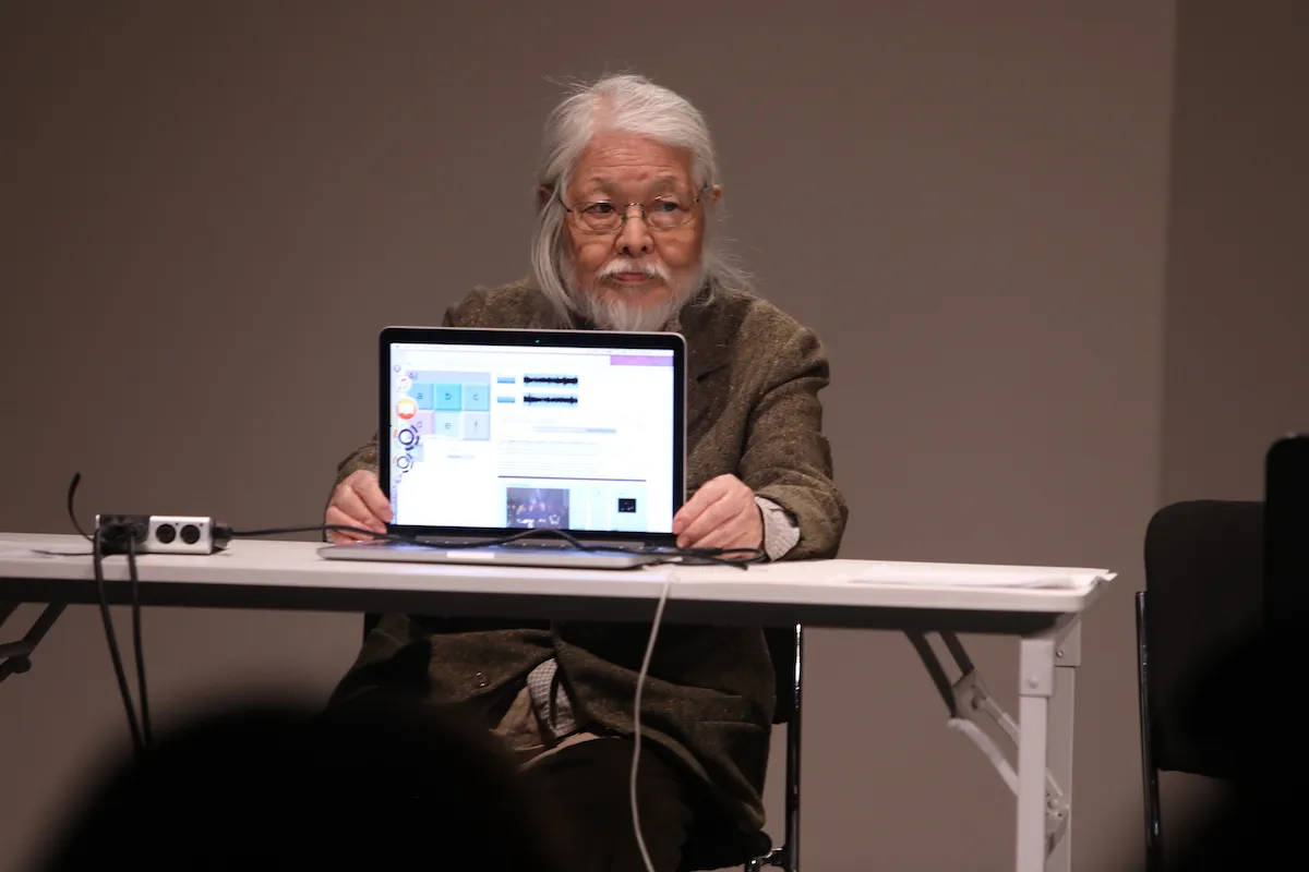 An elderly Asian man holding up a laptop screen to an audience.