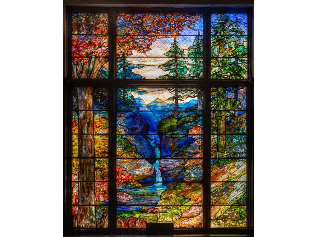 A monumental stained glass window showing a nature scene of a forrest, stream, and mountain the background.