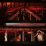 The upper levels of an industrial building, showing three performances moving their bodies on an upper level with a camera crew filming them on a level above.