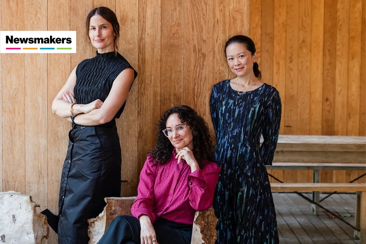 Three women, one seated, in front of a rustic yet refined wood panel wall.