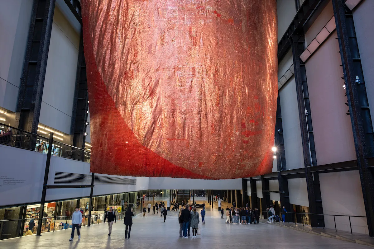 A large red-orange sculpture hanging down above people walking beneath it.