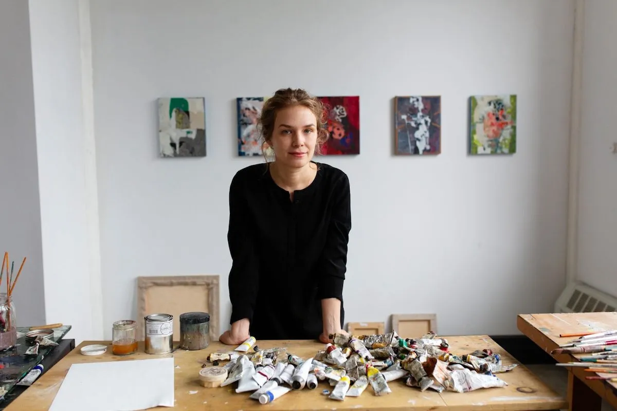 A woman standing at a table with tubes of paint on it. She stands in front of five abstract paintings.