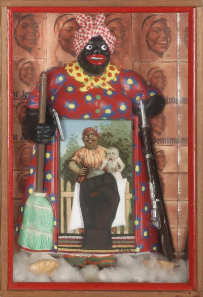 A sculpture of Aunt Jemima inset in a box. She holds a broom in one hand and a rifle in another. In front of her is a picture showing a mammy figure holding a white child.