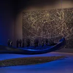 A blue-lit hanging canoe with hands sticking out of it in a gallery space with a painting on one wall.