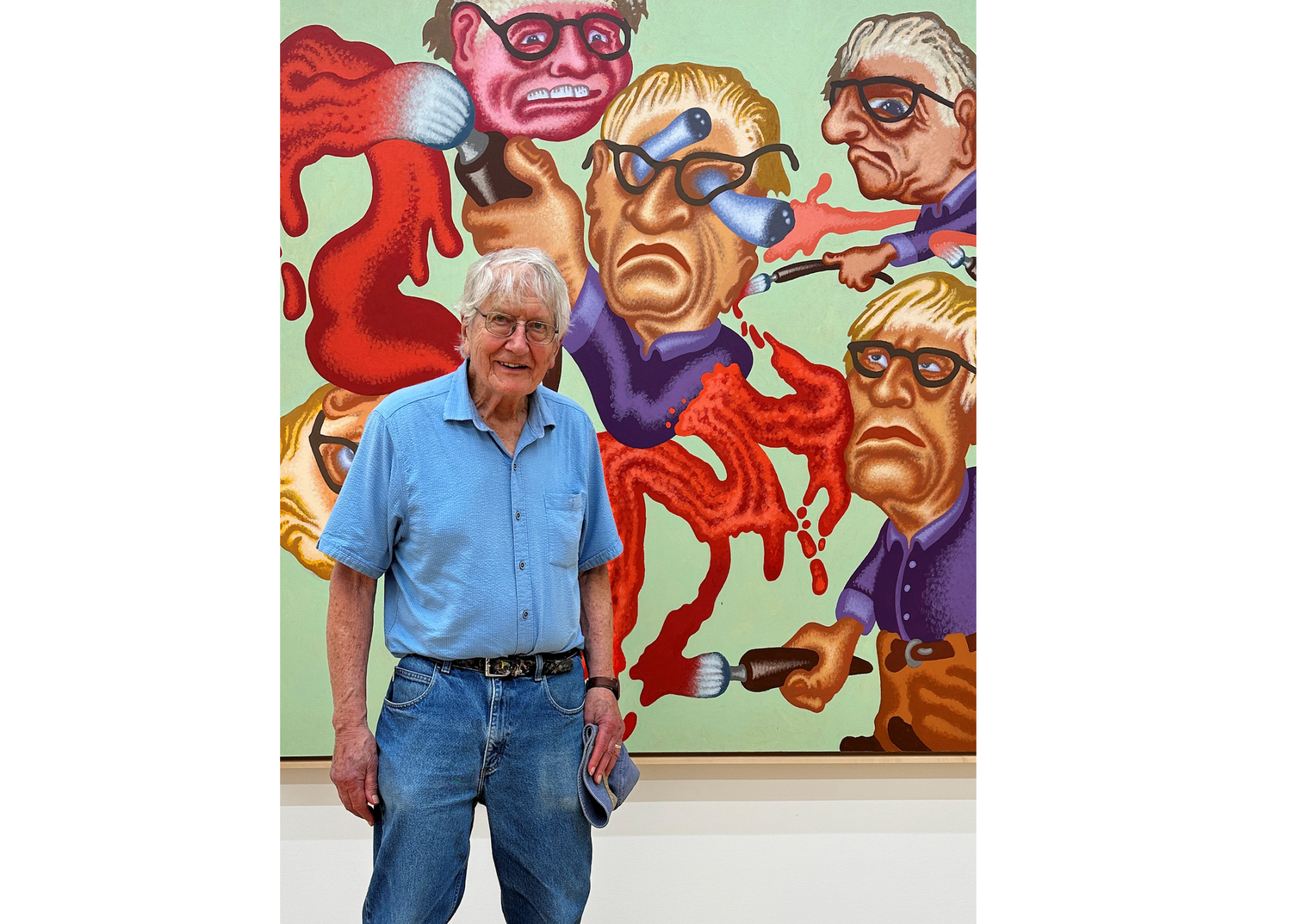 An older man with glasses smiles at the camera. Behind him is a pretty crazy painting of four similarly bespectacled men, one of whose eyes is popping out of his gourd.