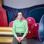 A man in a green shirt seated in front of a large painting of jewelry.