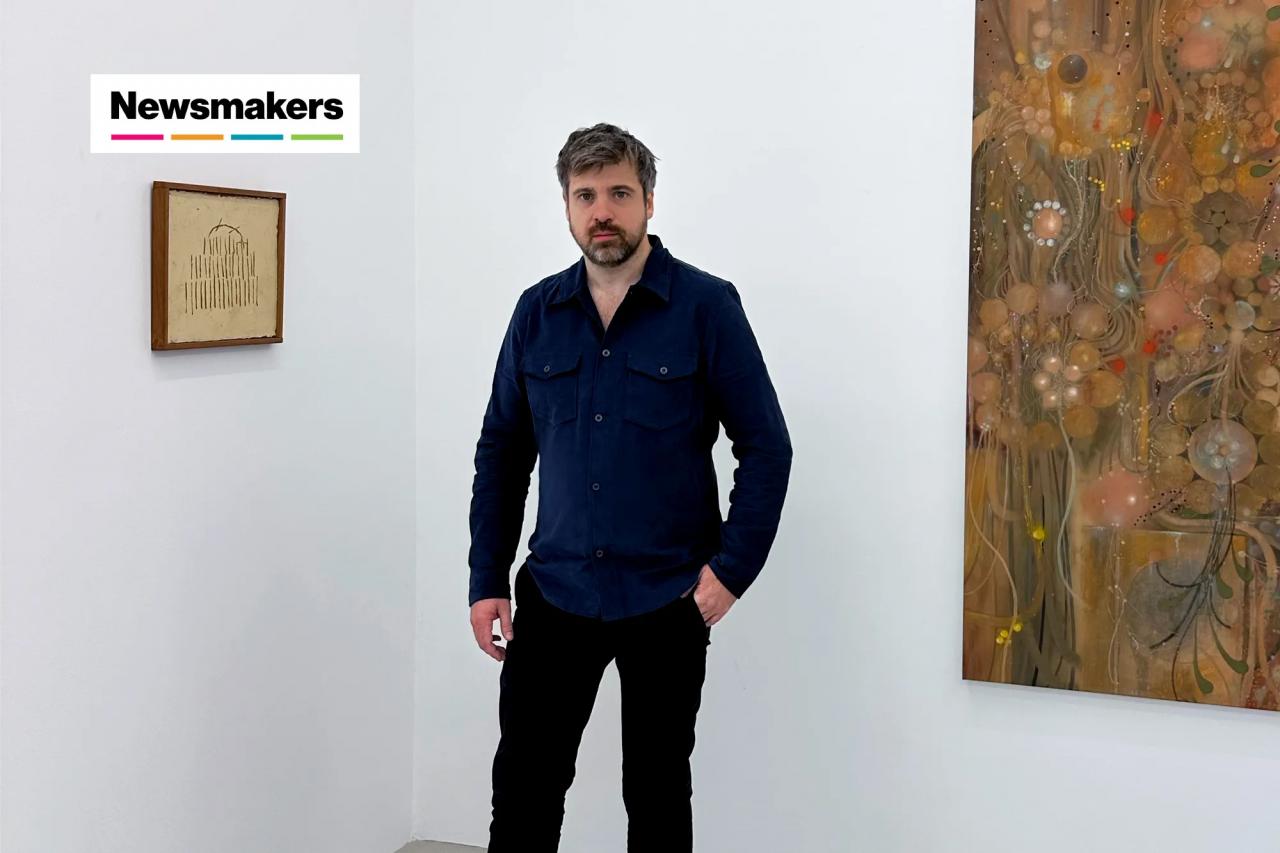 A man in a blue button down stands between two paintings.