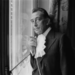A man in a suit with a long mustache standing beside a window.