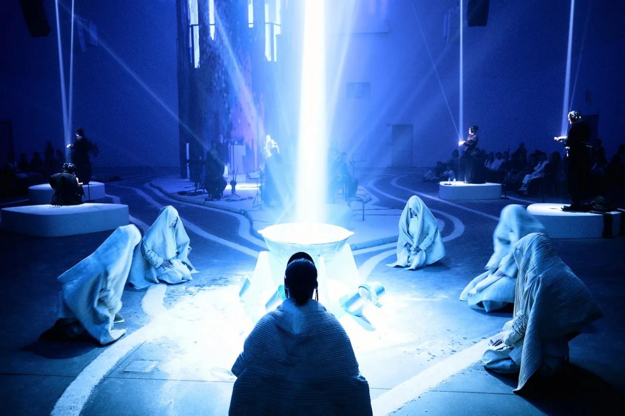 Several people in white robes centered around a cauldron emitting bright white light.