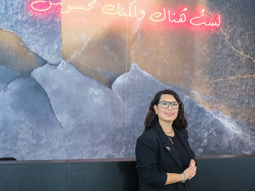 A woman with long dark hair, glasses, and a black suit stands in front of a blue and gray patterned wall with Arabic text in red neon above her head