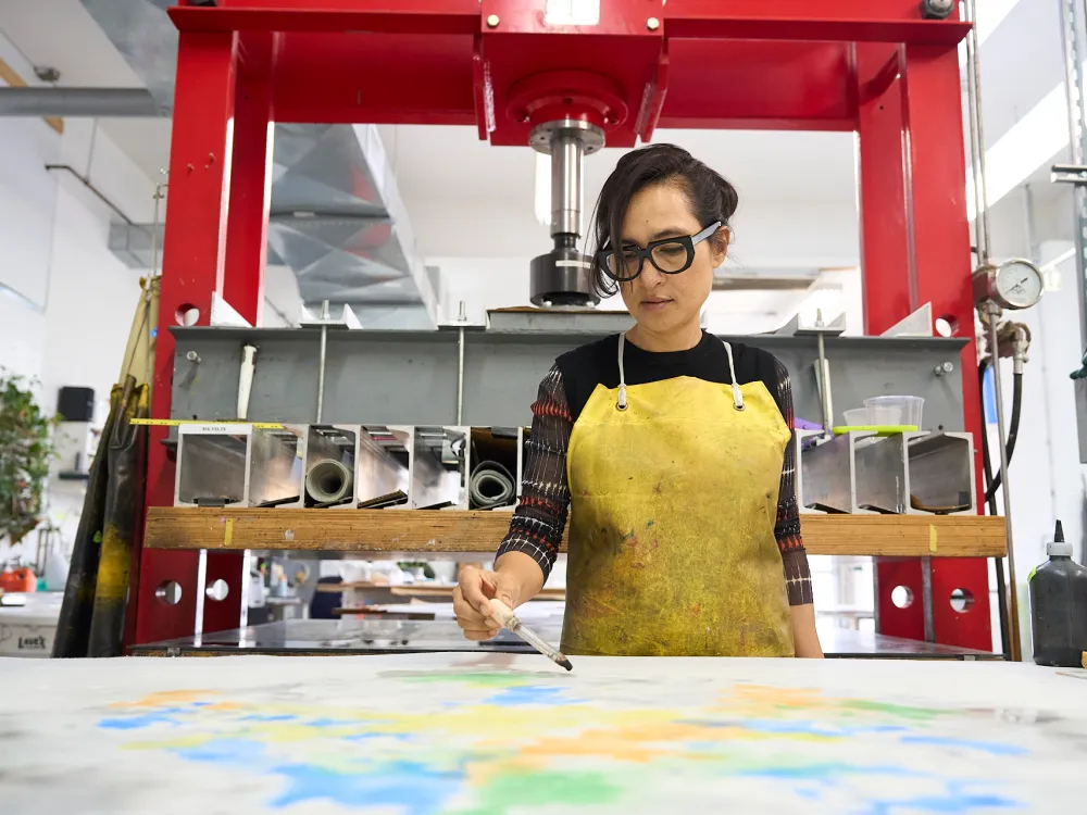 A woman with dark hair and dark glasses applies colored liquid to a work on paper.