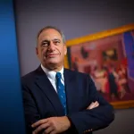 Portrait of John P. Axelrod, standing in a museum with a painting (out of focus) behind him.