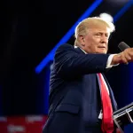 Donald Trump, in a suit with a red tie, stands at a microphone and points into the distance.