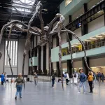 A sculpture of a giant spider in a big room with people walking underneath it.