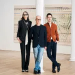 Two men and a woman stand near a white column in a white- walled gallery.