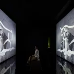 A woman watches a screen depicting a man painting.