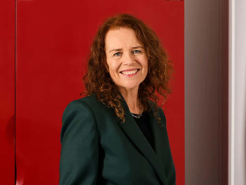 a smiling woman with wavy shoulder-length reddish hair, wearing a dark green blazer and standing in front of a red wall