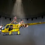 Four women climb and crawl over a helicopter that is suspended in the air.
