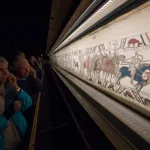 People looking at a tapestry showing people on horseback.