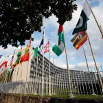 Flags of at least a dozen countries hang on flagpoles in front of a curving modernist building of perhaps eight or night stories.