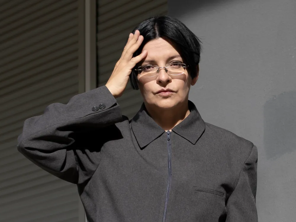 Portrait of Henrike Naumann in a grey jacket with a zipper and collar holding one hand to her head.