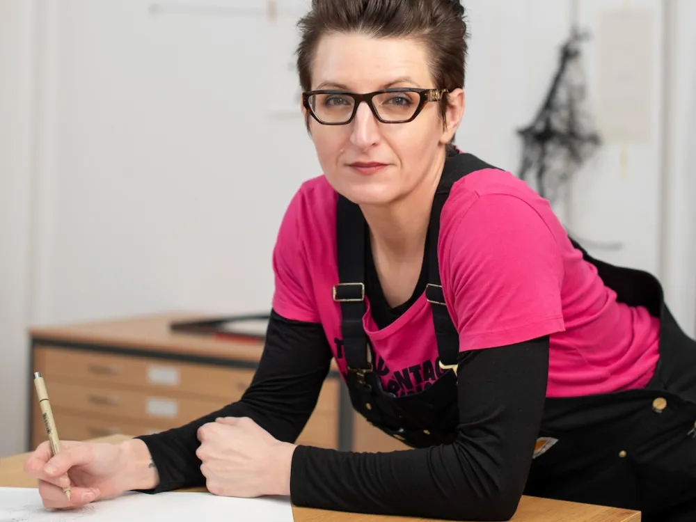A woman with glasses and a pink shirt holds a drawing pen and leans on a table.