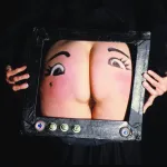 A taped frame with buttons encloses a but with eyes painted on it. It looks like a TV.