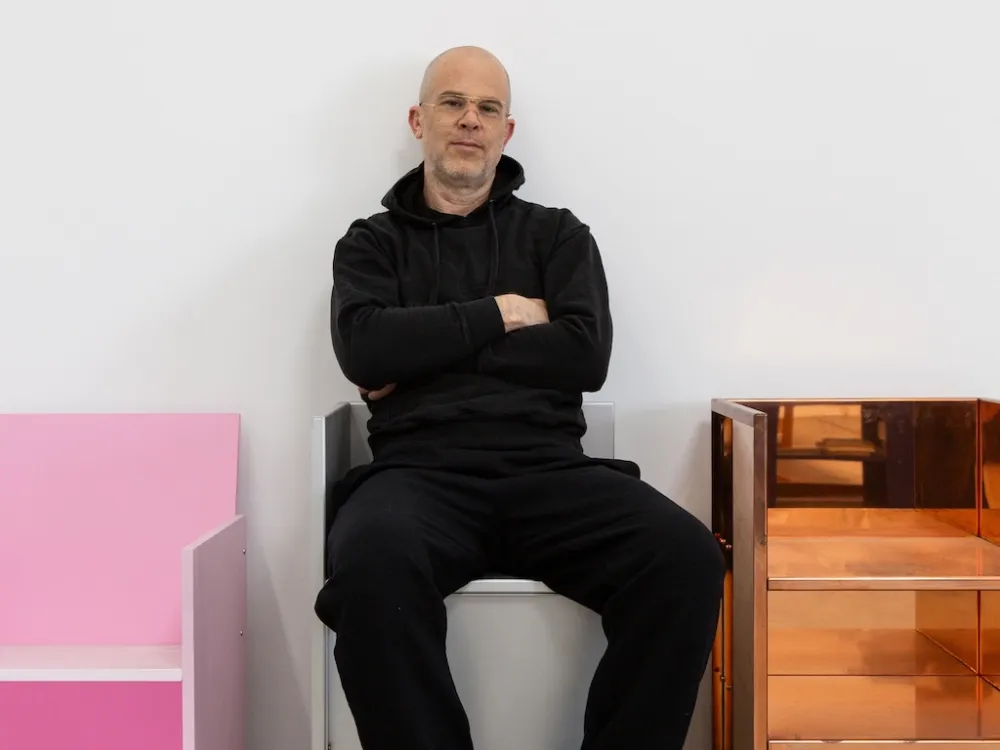A man in dressing black sitting on a white chair between a pink chair and a brass chair.