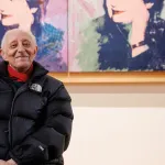 A man in a black puffer jacket sitting in front of a painting by Andy Warhol.
