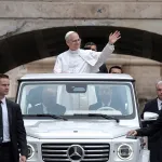A man in a white outfit waves from a car.