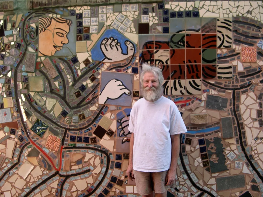 An elderly man with white hair and beard, artist Isaiah Zagar, poses smiling in front of a mosaic artwork he created