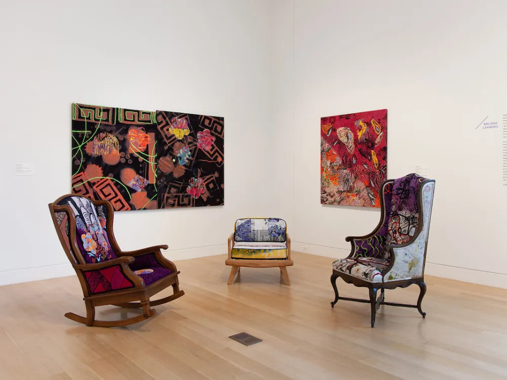 An installation composed of a rocking chair and a high-backed chair set before two paintings.