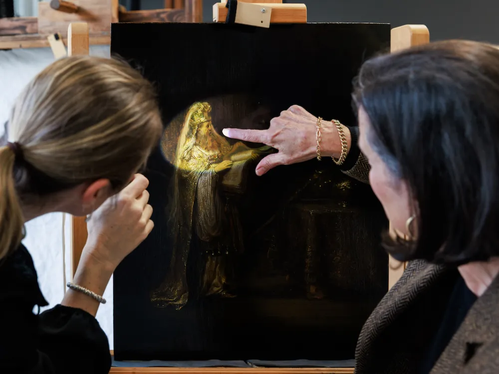 Two women pointing at a canvas painted with an image of a man in golden robes illuminated by a spotlight.