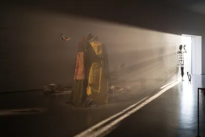 A darkened gallery with light pouring in that illuminates a metal sculpture of a person with an armature for a body.