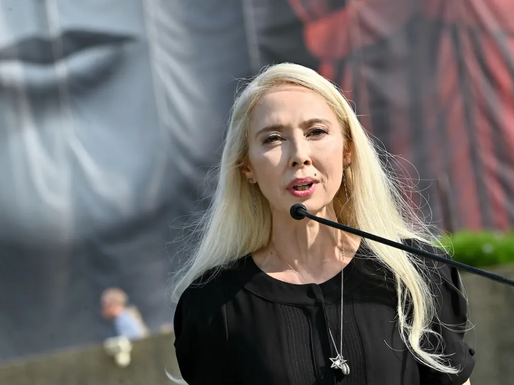 Portrait of Melissa Chiu speaking into a microphone before a large painting of a person