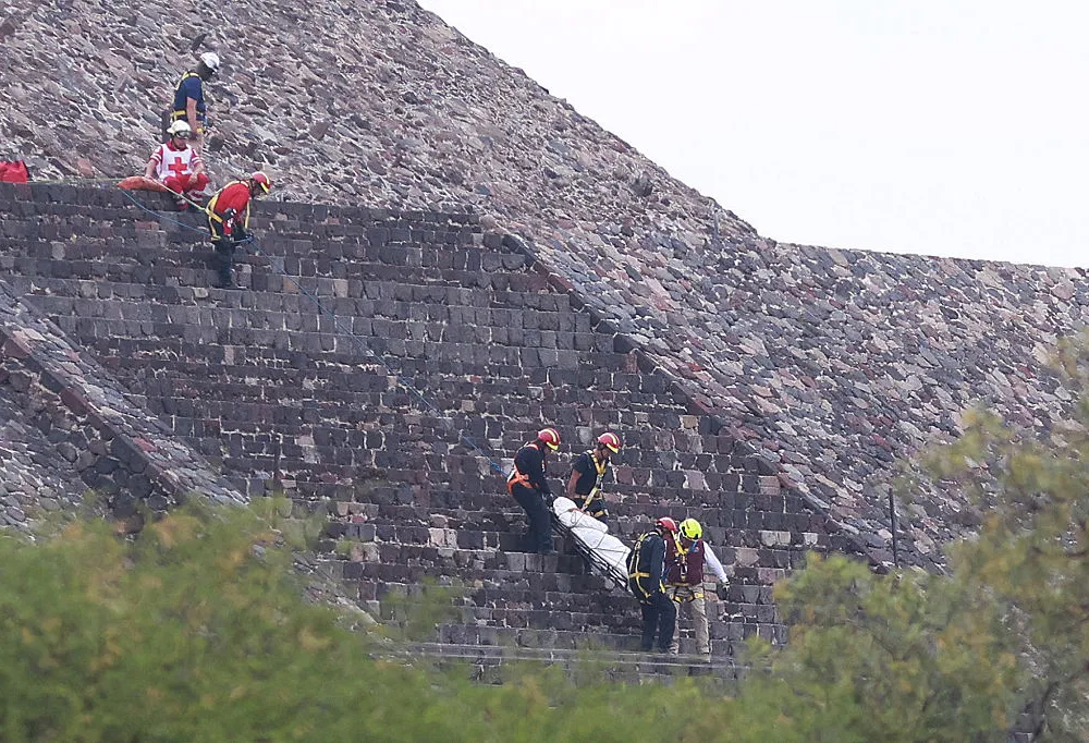 A body is carried on a stretcher down the steps of a grey stone pyramid.