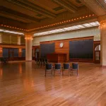An empty room with two rows of chairs in it. Heavy columns with lights strung around them hold up an ornate ceiling.