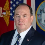 A white middle-aged man, John Phelan, in a suit and tie and with a US flag lapel pin, sits for a portrait in front of a U.S. flag and other flags