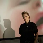 A woman in a black shirt stands in front of a film screen showing a baby face.