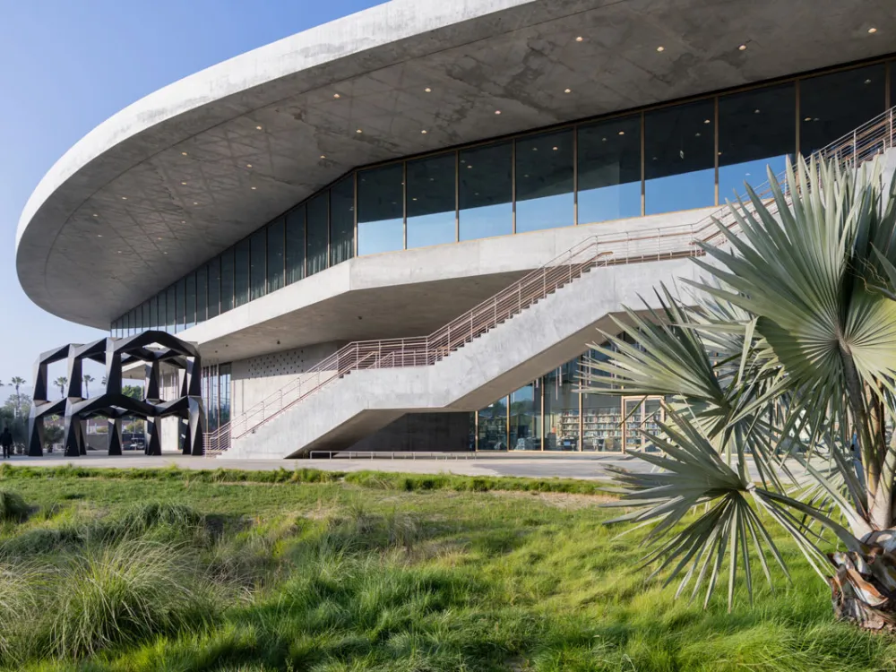 A curved concrete building with an abstract black sculpture at left. In the foreground are plants.