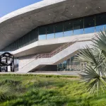 A curved concrete building with an abstract black sculpture at left. In the foreground are plants.