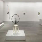 White walls of a gallery filled with ready-made works including a upside-down bicycle wheel on a stool and a shovel hanging from the ceiling.