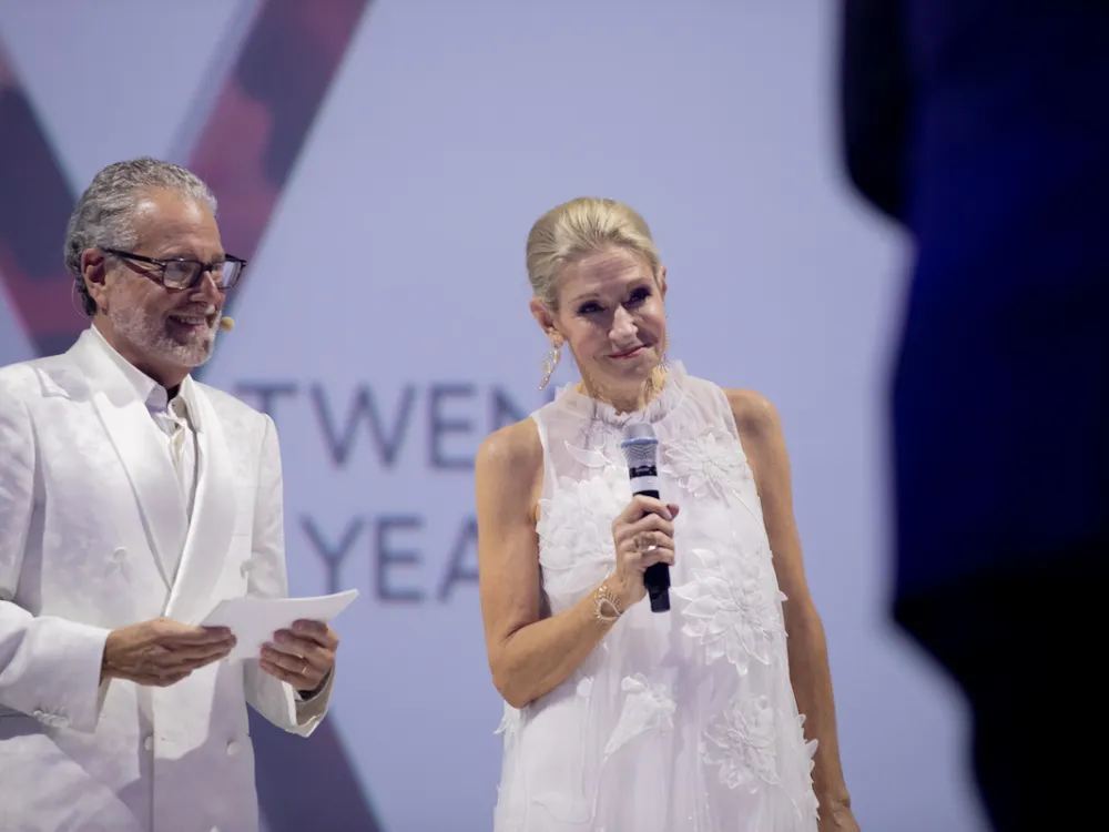 A man and a woman, both wearing white formal wear, on a stage. The main holds papers, while the woman holds a microphone.