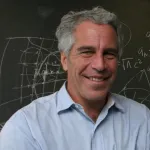 A white man with salt and pepper hair smiling in a blue shirt. He stands in front of a blackboard with mathematical equations scrawled on it.