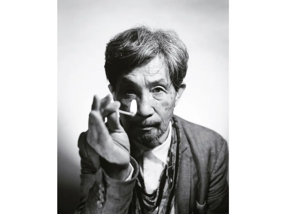 Black and white portrait of Gozo Yoshimasu, who holds a lit match in front of his face.