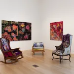 An installation composed of a rocking chair and a high-backed chair set before two paintings.